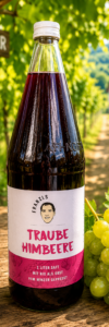 Produktfoto Traube Himbeer Heuriger Eine Glasflasche mit Traube-Himbeersaft von „Franzls Fruchtsäfte“ steht im Fokus auf einem rustikalen Holztisch in einem sonnendurchfluteten Heurigen-Weingarten. Im weich gezeichneten Hintergrund schimmern grüne Weinreben im warmen Nachmittagslicht. Neben der Flasche liegt eine frische Rebe grüner Trauben.